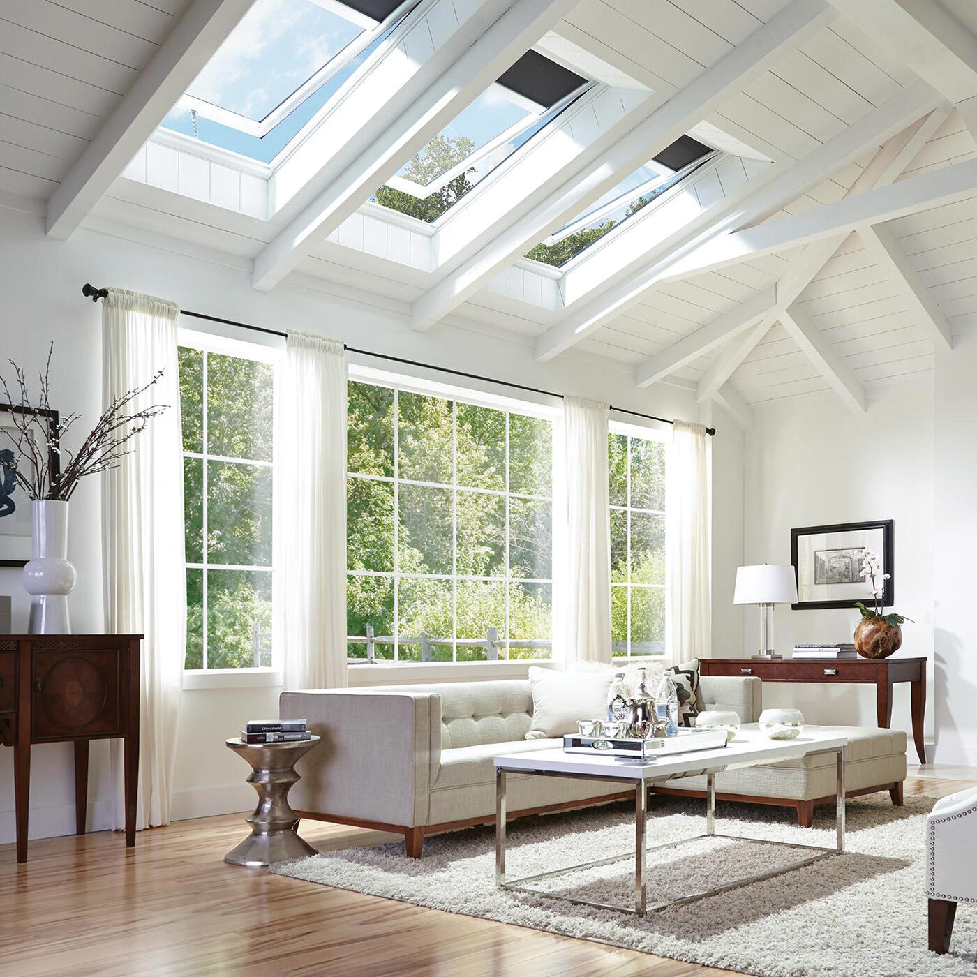 Bright living room with large windows, white walls, white sofa, glass coffee table, and skylights in the ceiling showing blue sky and trees.